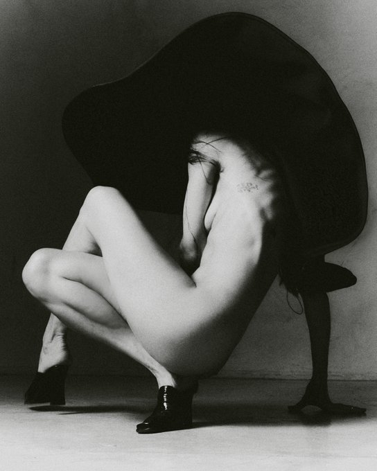 A striking black and white image captures a person in a deep squat, draped by an oversized hat, with a subtle tattoo visible on their back. This artistic shot is shared in relation to the @objktcom platform, hinting at a digital art or NFT context.