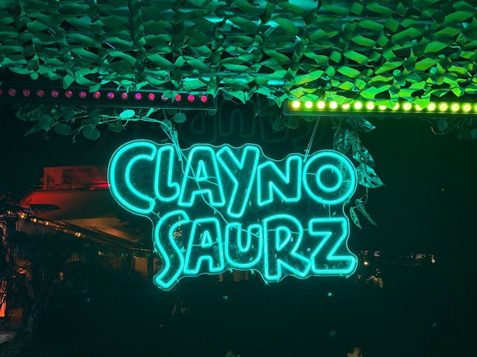 A glowing cyan neon sign reading 'CLAYNO SAURZ' mounted beneath a ceiling of green leaves and colorful LED strip lights in a dark setting.
