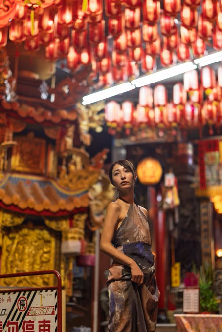 A woman wearing a sheer halter dress stands in front of an ornate Chinese temple decorated with numerous glowing red lanterns hanging overhead.
