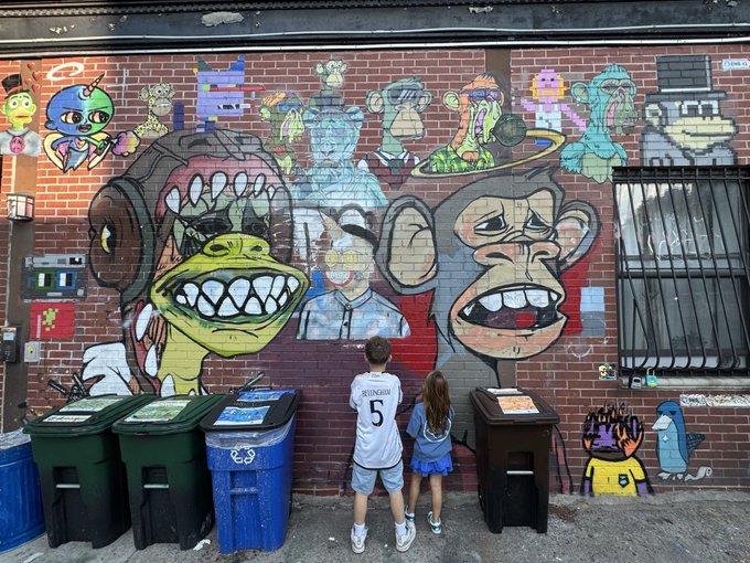 Two children stand before a large brick wall covered in colorful graffiti art featuring Bored Ape-style characters and various cartoon figures, with trash bins in the foreground.