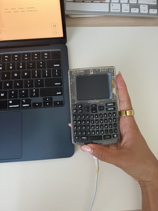A hand holds a transparent Coldcard hardware wallet connected to a laptop displaying its home screen, with a keyboard visible in the background. The image illustrates the post's message about a 2021 firmware vulnerability in Coldcard devices, prompting the author to abandon their long-time recommendation and move their assets elsewhere.