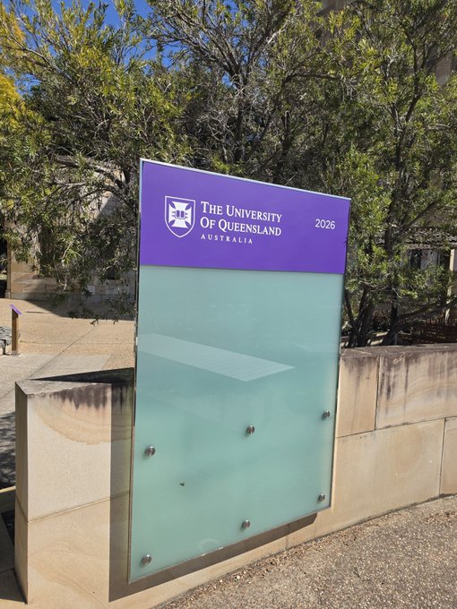Outdoor sign for 'The University of Queensland Australia 2026' mounted on a stone wall, with a frosted glass panel below and trees in the background.