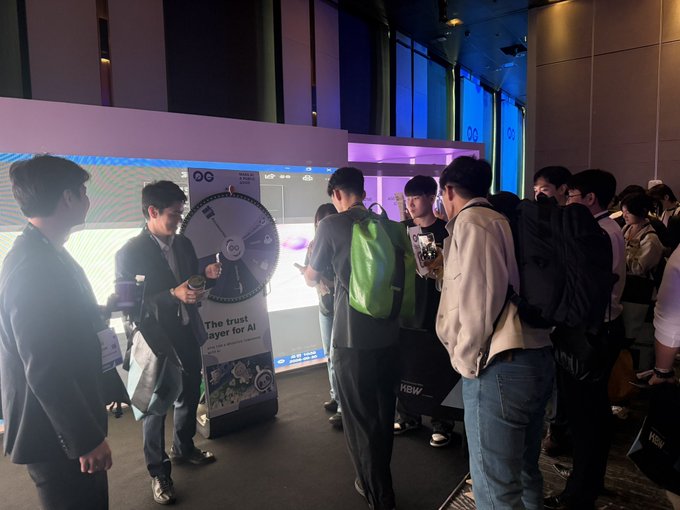 Photo from a tech conference or event showing people gathered around a booth with a spinning prize wheel labeled 'The trust layer for AI' by 'QG'.