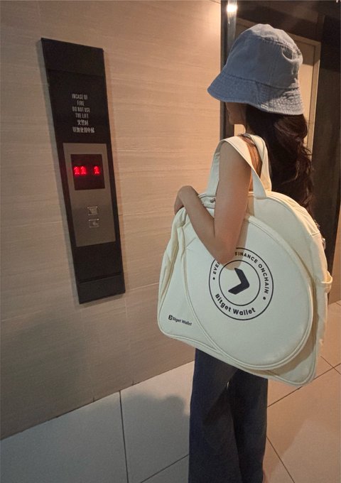 A woman in a denim bucket hat carries a large white duffel bag with the 'Bidget Wallet' logo while waiting for an elevator.