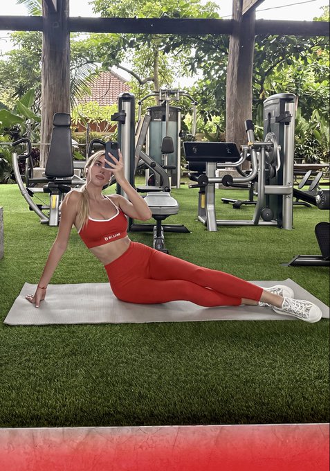 A woman in a red sports bra and leggings takes a mirror selfie on a yoga mat at an outdoor gym with green artificial turf and exercise equipment.