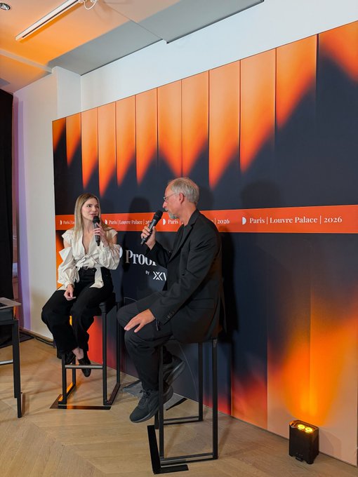 The image shows a man and woman sitting behind a microphone, likely participating in an event or discussion. The man appears to be wearing a suit while the woman has her hair styled in pigtails. They are both holding microphones, suggesting they might be hosting a talk show or panel discussion. The background features a large orange wall with black text on it, which could be related to the event or the topic being discussed by the hosts.