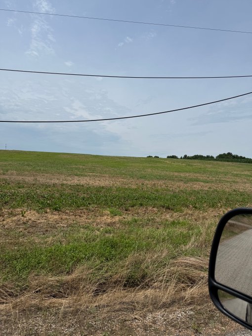 A view from a moving vehicle shows a field of cut grass under a partly cloudy sky, with power lines overhead and a side mirror visible in the lower right corner.
