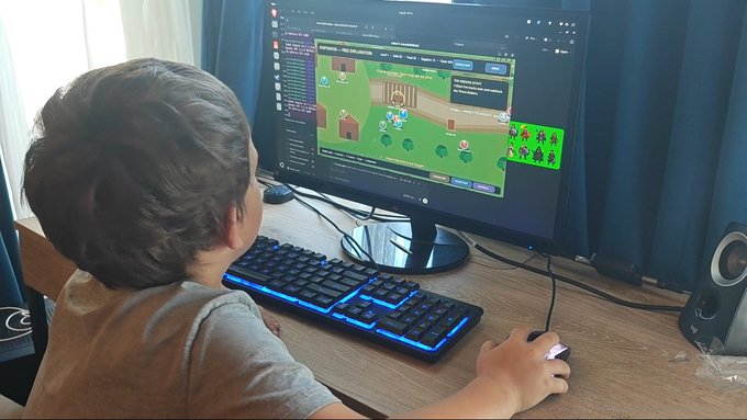 A young boy wearing headphones sitting at a computer desk, using a blue-backlit keyboard to play what appears to be a farming or building game on his monitor.
