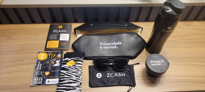 A flat-lay display of ZCASH branded merchandise including a black water bottle, sunglasses, socks, a toiletry bag reading "Privacidade é normal," and stickers on a wooden table.