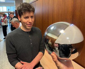 The image shows a man holding up a silver ball with a reflective surface, possibly a globe or a mirror ball. The scene takes place in front of a wooden wall, which could be part of an office building or another type of establishment. There are several other people present in the background, but they are not the main focus of the image. The man holding up the ball appears to be the center of attention as he poses with it for the camera.