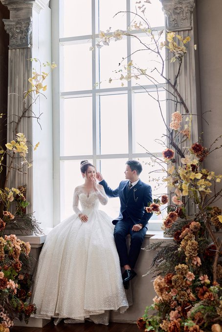 A bride in a white ball gown and a groom in a dark suit pose together by a large window, framed by elaborate floral arrangements with yellow and orange blooms.