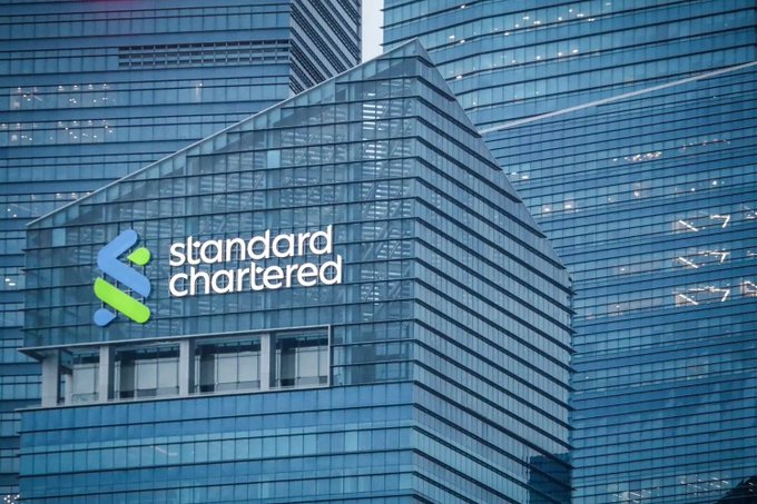 A close-up photograph of a glass skyscraper displaying the 'standard chartered' bank logo in blue and green.