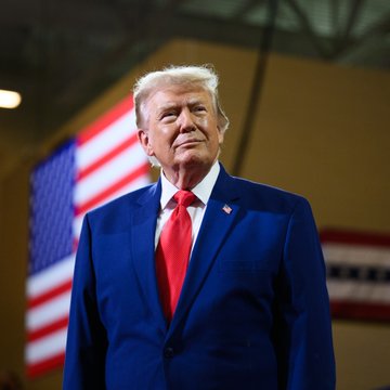 President Donald Trump stands at a podium in a navy blue suit with a red tie, with American flags displayed behind him, as he appears during an interview discussing the United States' significant investments and leadership in artificial intelligence.