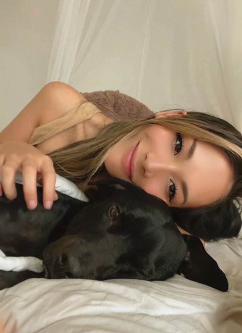 A woman with long brown hair lies on a bed next to a black dog, looking at the camera.