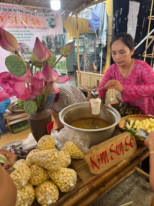 The image shows a woman pouring soup from a large pot into a bowl, while another person watches her closely. The scene takes place at an outdoor market or food stall where there are several bags of popcorn displayed nearby. A sign for "KIM SONGS" is also present in the scene.
