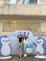 Two women taking a picture in front of a 'PiPi & Kii' themed penguin photo stand at a theme park.