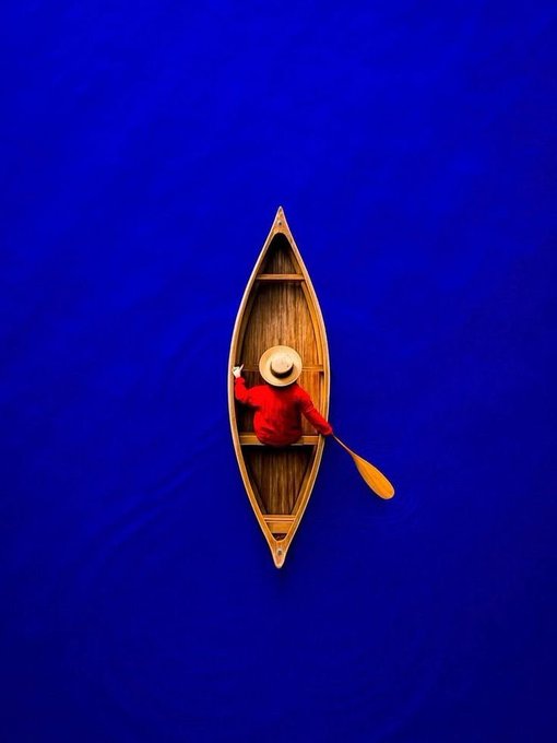 An aerial view of a person wearing a red shirt and straw hat paddling a wooden canoe on deep blue water.