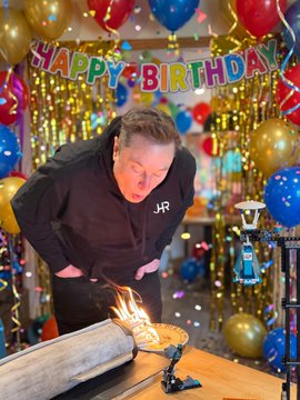 Elon Musk blows out candles on a birthday cake, with a "HAPPY BIRTHDAY" banner and festive balloons decorating the background.