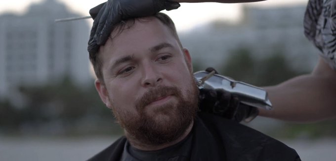 A bearded man with tears in his eyes receiving a haircut outdoors, with someone wearing black gloves using clippers to shave the side of his head in what appears to be an emotional moment.