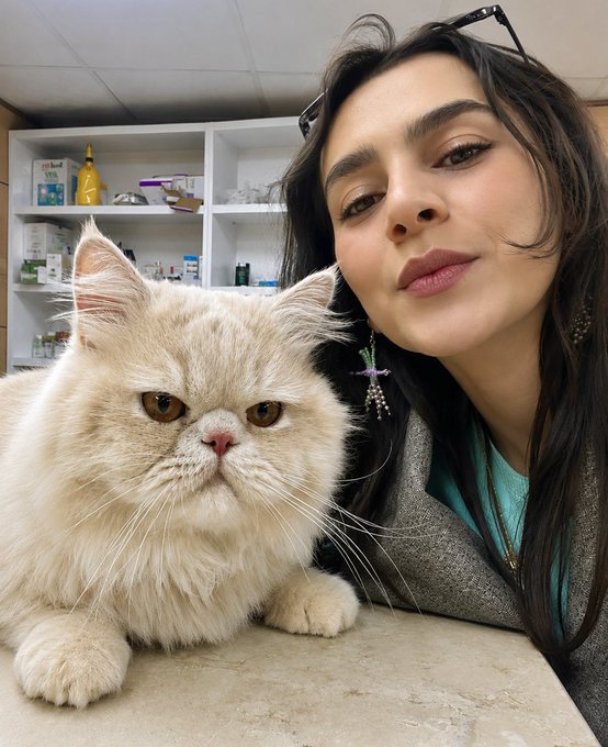 A woman poses for a selfie with a fluffy cream-colored cat in what appears to be a veterinary clinic, with medical supplies visible on shelves in the background.