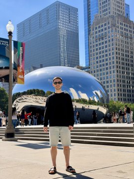 Image shows a man in a black shirt, khaki shorts, and sunglasses standing in front of the Cloud Gate (Chicago Bean) sculpture in Millennium Park.
