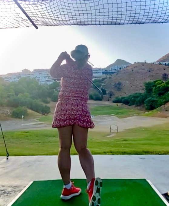A woman in a pink patterned dress and orange golf shoes swings a club at a golf driving range with hills in the background.