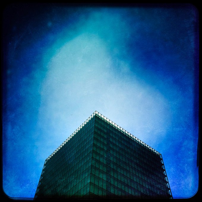 A glass skyscraper's corner rises into a vivid blue sky, its geometric peak capped with a railing and surrounded by soft, cloud-like light—visually echoing the post's "Glass Crown" imagery and "sharp blue geometry suspended between light and stillness."