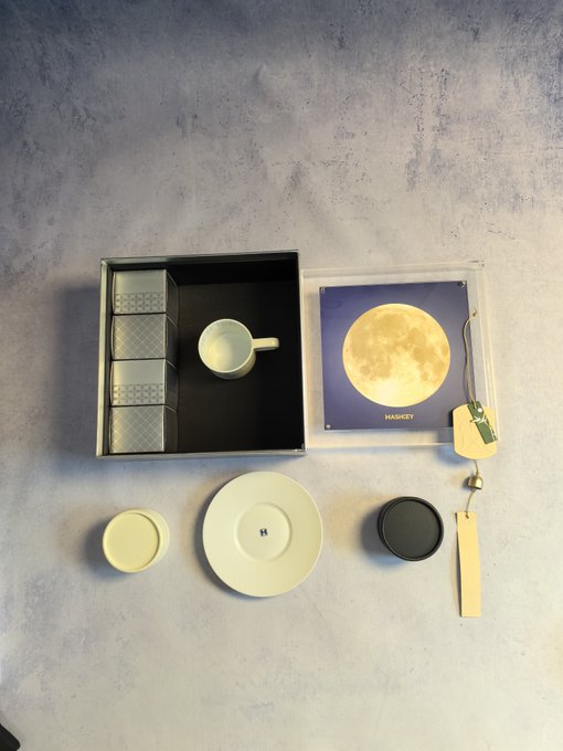 An overhead view of a neatly arranged product set on a textured surface, featuring a black box with metal tins, a white mug, a plate with an 'H' logo, a moon-themed card labeled 'HAMIDEY', a black lid, and a strap.