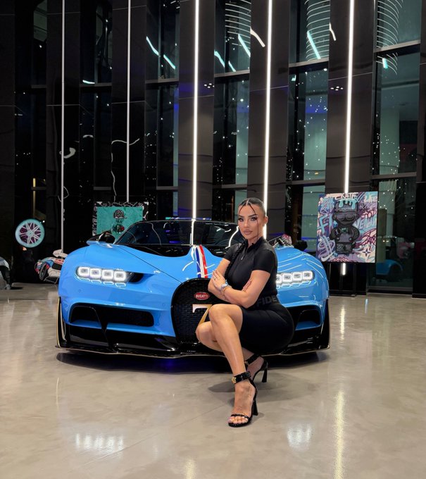 A woman in a black dress poses in front of a blue Bugatti hypercar inside a luxurious showroom with vertical light strips and graffiti-style artwork.