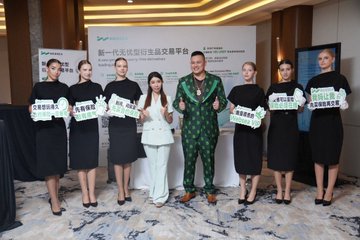 A group photo of seven people at a press event, with a man in a green patterned suit standing in the center alongside women in black dresses holding promotional signs, with Chinese text on the backdrop reading '新一代功效白芸豆革平台' (New Generation Effective White Kidney Bean Platform).