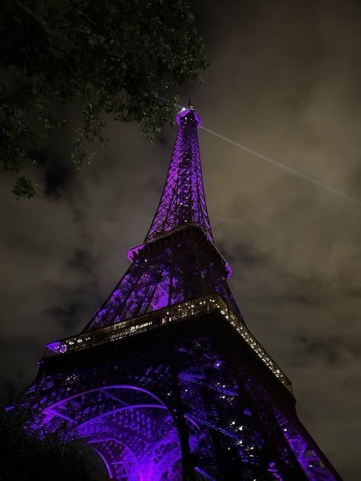 The Eiffel Tower is illuminated in purple light at night against a cloudy sky.