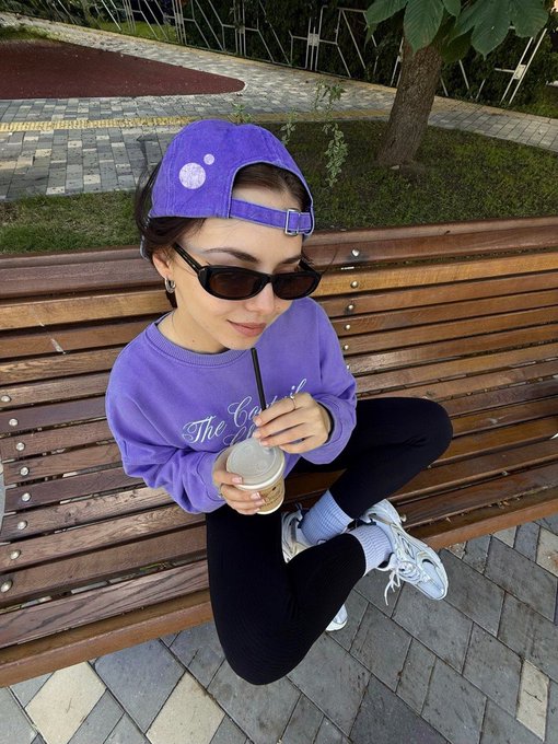 A young woman wearing a purple cap, sunglasses, and a purple 'The Cookie' sweatshirt sits cross-legged on a wooden bench holding an iced coffee drink.