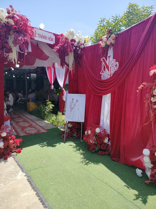 A Vietnamese wedding venue entrance decorated in red and white floral arrangements with a red curtain backdrop displaying 'LỄ THÀNH HÔN' and a wedding photo of the couple.
