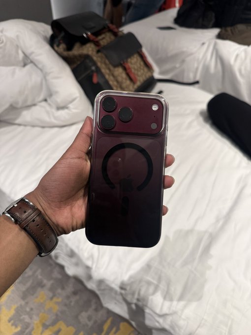 A hand wearing a brown leather watch strap holds up a burgundy/dark red iPhone (likely an iPhone 15 Pro) encased in a clear cover with a MagSafe ring visible on the back. In the background, a Gucci monogram messenger bag rests on a white bed, suggesting the person is freshly arrived and prepped for the Token2049 crypto conference.