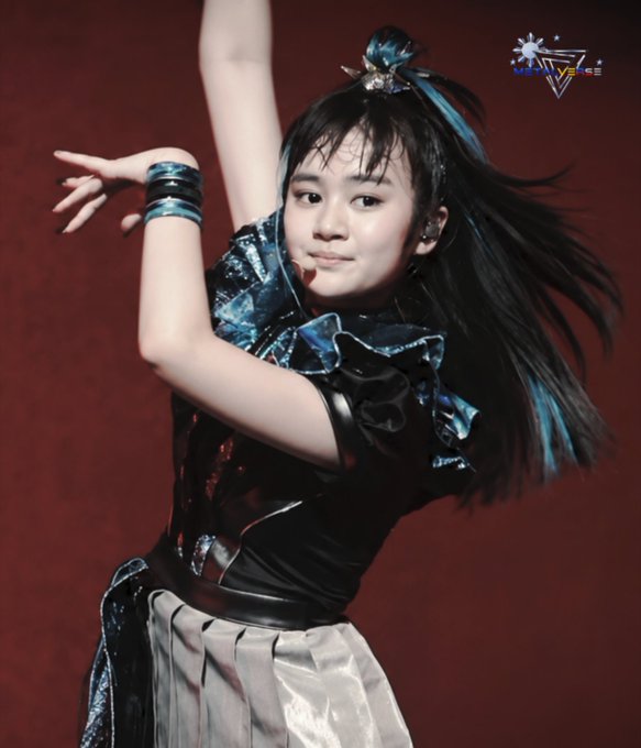 A performer on stage in a blue and black outfit with teal arm bands, posing dynamically with one arm raised against a red backdrop, with a logo visible in the corner.