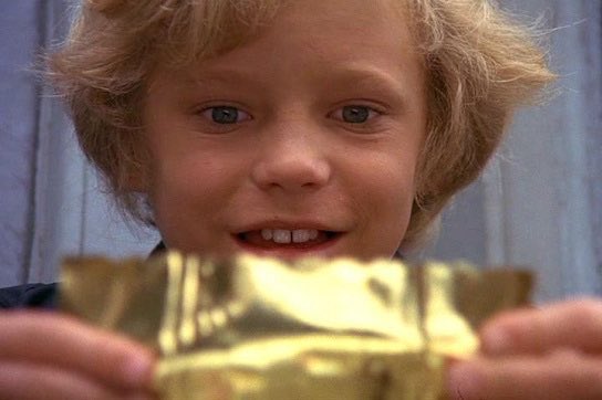 A young blonde boy smiles while holding up a gold foil package, with a cut on his lip visible.