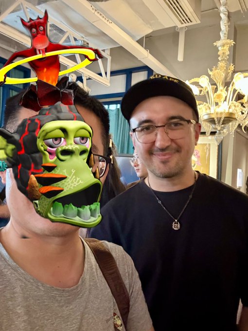 A man wearing glasses, a black cap, and a peace sign necklace poses beside another person whose face is obscured by a digital overlay of a Bored Ape Yacht Club (MAYC) NFT with a red devil-like figure above it, set in an upscale interior with an ornate chandelier—visually representing the in-person meetup mentioned in the caption with @mfigge in Shanghai.