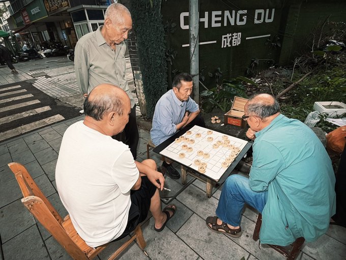 Four elderly men gather on a sidewalk in Chengdu (with "CHENG DU" and "成都" visible on the wall behind them), playing a board game around a small table as another man stands watching. The scene captures a quiet, communal moment of leisure among seniors, though it doesn't directly relate to the accompanying text about collecting Mid-Autumn Festival merchandise.