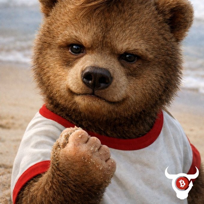 A photorealistic image of an adorable bear cub wearing a white and red shirt, sitting on a beach with sand on its paw, featuring a Bitcoin bull logo watermark in the corner.