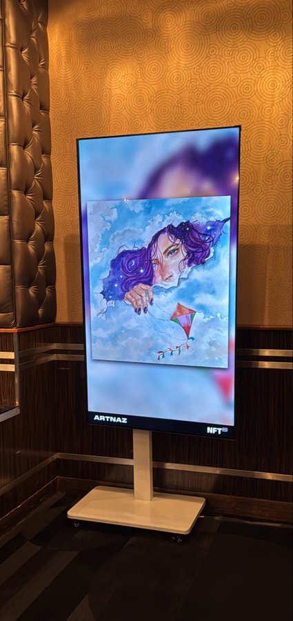 A digital kiosk displaying an artwork of a purple-haired woman reaching for a kite in clouds, labeled 'ARTNAZ NFT' at the bottom.