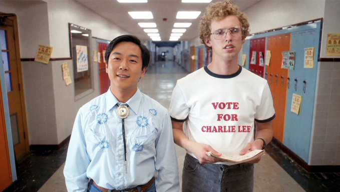 Two young men stand in a school hallway with colorful lockers, the taller one on the right wearing a white t-shirt reading "VOTE FOR CHARLIE LEE" and holding a clipboard, while his shorter companion on the left wears a blue embroidered shirt with a bolo tie. The image humorously illustrates the post's slogan "A vote for Lee is a vote for me," with the shorter friend visibly endorsing his taller counterpart's campaign by literally wearing his candidate's name.