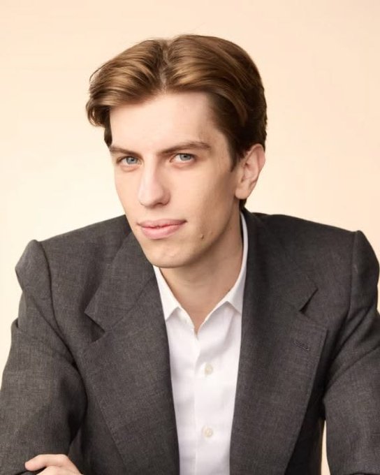 A portrait of a young man with light brown hair wearing a gray suit jacket over a white collared shirt, posing against a plain beige background.