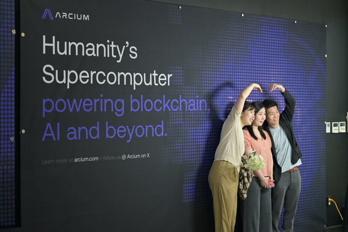 The image features three people posing together against a backdrop of a large banner that reads "Humany's Supercomputer powering blockchain AI and beyond." The group appears to be enjoying themselves, with one person holding up their hands as if they are making a peace sign or doing the "shaka" gesture. They seem to be at an event related to cryptocurrency or blockchain technology, possibly celebrating a recent success or milestone in the industry.