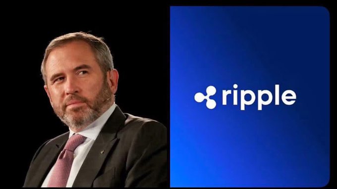 The image features a man wearing a suit, sitting next to a blue sign that says "ripple" written in white letters. The man appears to be looking at the camera with a serious expression. The background of the image is black, which contrasts with the blue color of the sign and makes it stand out more prominently.