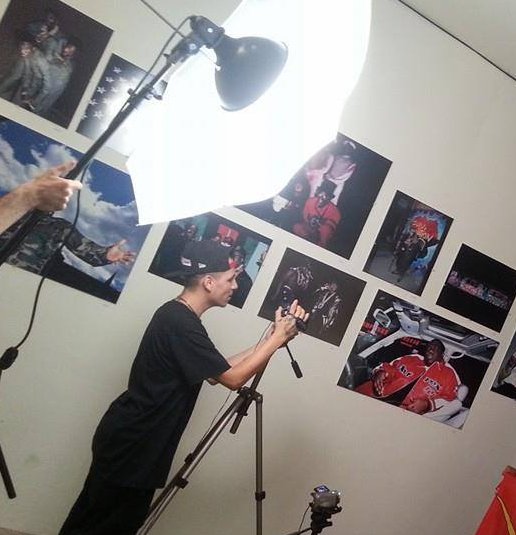 A photographer stands behind a tripod in a studio setting, preparing his camera beneath a large lighting umbrella while framed photographs decorate the wall behind him.