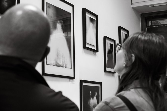 A black-and-white photograph of two people viewing framed artwork displayed on a gallery wall.