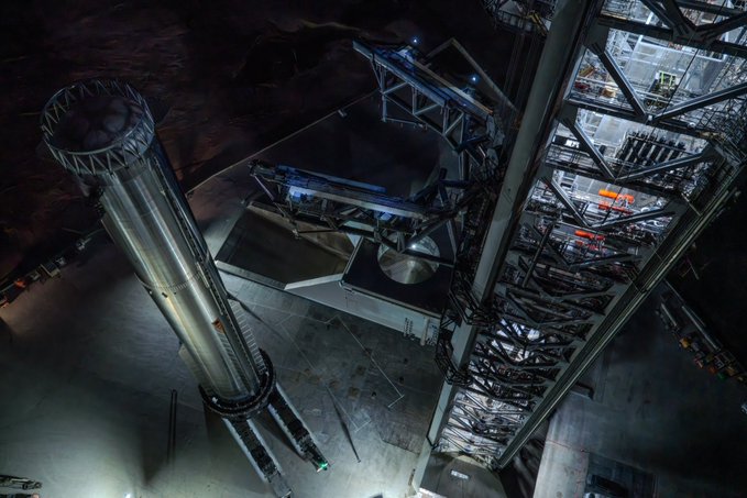 An aerial nighttime view of SpaceX's Starship rocket on the launch pad, showing its tall silver booster and the launch tower's mechanical arms.