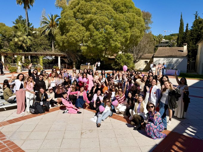 A large group photo of people posing together outdoors on a sunny day, with trees and a building in the background; pink balloons are visible in the crowd.