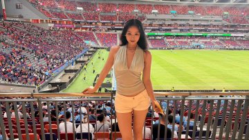 A young woman poses in the stands of a packed football stadium, smiling at the camera while enjoying her first live match, as mentioned in the post thanking OKX for the invitation. The vibrant green pitch and rows of cheering fans create an electric matchday atmosphere behind her.