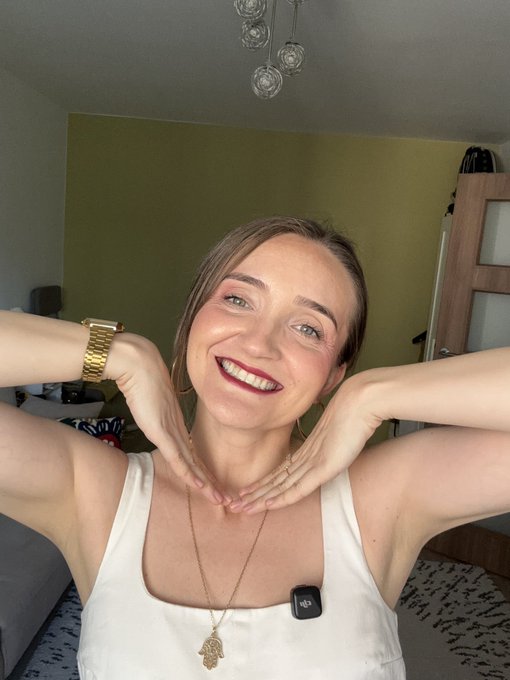 A smiling woman with blonde hair framing her face with her hands, wearing a white tank top, gold watch, and Hamsa necklace, with a small DJI mic clipped to her shirt in a room with green walls and decorative hanging lights.
